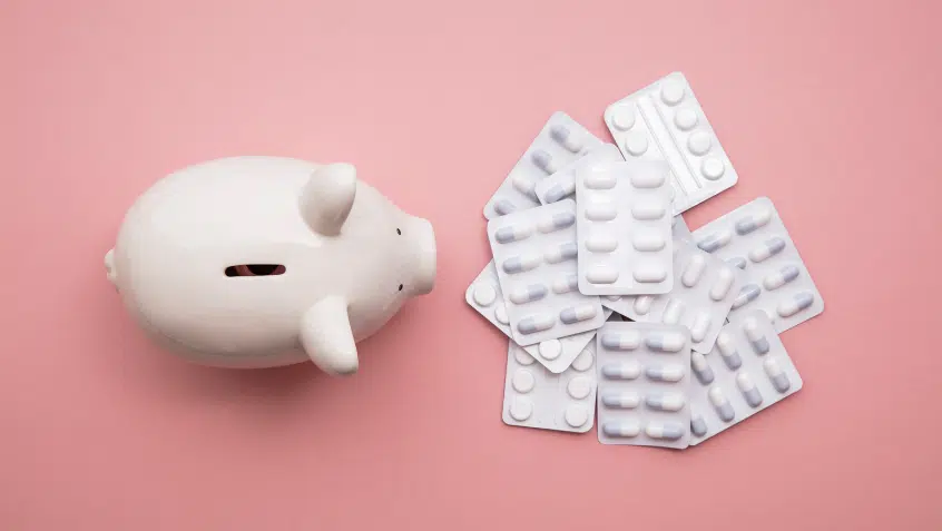 On a pink background, a photo taken from above shows a piggy bank beside a pile of pills